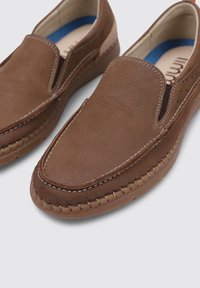 Zapatos marrones de cuero tipo slip-on con una superficie texturizada, costuras blancas y una suela de goma flexible. Presentan un forro interior azul.