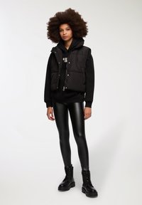 Gilet imbottito nero con cappuccio, indossato sopra un hoodie nero. Il modello indossa leggings neri lucidi e stivali neri con lacci. Design semplice e moderno.