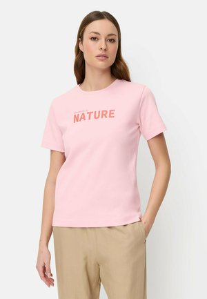 Femme portant un t-shirt rose clair avec le texte « renforcée par la NATURE », un pantalon beige, debout avec une main dans la poche sur un fond blanc.