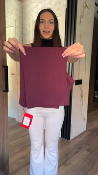Femme aux longs cheveux foncés tenant un vêtement violet plié avec une étiquette de prix rouge dans une pièce moderne avec des accents en bois et marbre.