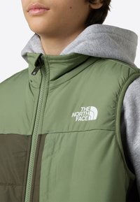 Vadderad ärmlös väst i ljusgrön med mörkgrön panel, med dragkedja framtill och vitt logotyp på bröstet. Grå huvtröja syns.