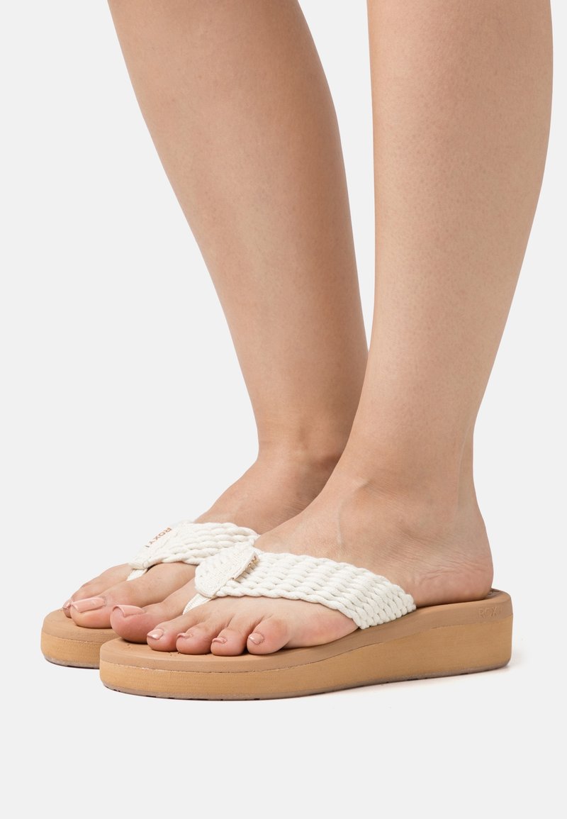 Des sandales slip-on blanches tissées avec un dessus tressé et une semelle épaisse couleur tan. Le design comprend une semelle intérieure rembourrée pour le confort.