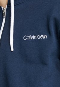 Sudadera con cremallera en color marino, hecha de una tela suave, con un cordón blanco, un logo bordado de "CalvinKlein" en el pecho y una textura suave.