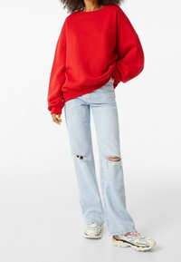 Sweat-shirt rouge surdimensionné, jean bleu clair déchiré avec ourlets effilochés, et baskets épaisses multicolores. Texture douce, coupe décontractée.