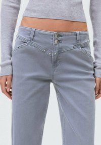 Sive denim hlače z širokimi nogavicami, dvema kovinskima gumboma in okrasnimi žebljički ob žepih in šivih. Gladka tekstura.