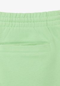 Un pantalon de jogging vert clair présente une taille élastique et une poche arrière avec un accent cousu. Le tissu est lisse et décontracté en apparence.
