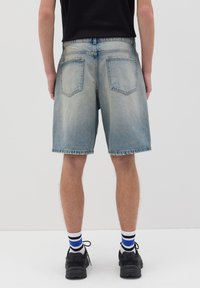 Pantalones cortos de mezclilla azul claro con un acabado desgastado, con dobladillo sin acabar y bolsillos traseros, combinados con zapatillas negras y calcetines rayados.