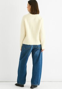Woman with straight brown hair wearing a cream fuzzy sweater, loose blue jeans, and black shoes standing facing a white wall.