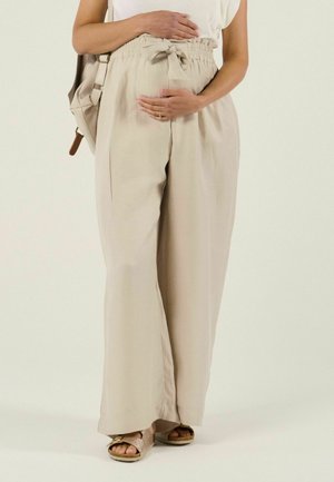 Personne enceinte portant un pantalon beige à jambes larges et des sandales, tenant son ventre avec les deux mains, portant un sac à dos beige sur l'épaule.