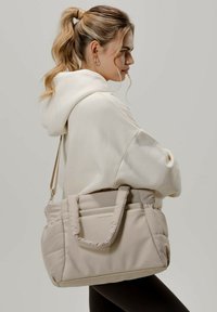 Sac fourre-tout beige rembourré avec deux poignées et une bandoulière réglable. Tissu lisse, couture discrète et petit logo à l'avant.