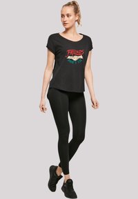 Schwarzes T-Shirt mit grafischem Design, das rote, grüne und weiße Schriftzüge zeigt. Kombiniert mit schwarzen Leggings und schwarzen Turnschuhen.