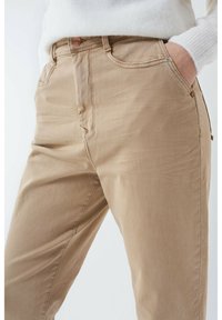 Pantalones de algodón beige con corte recto, que cuentan con bolsillos frontales, detalles de costura sutil y un cierre de botón metálico.