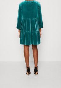 Tyrkysové sametové šaty s vícestupňovým designem, rukávy tři čtvrtě, a kulatým výstřihem, spárované s černými vysokými podpatky se zavazováním.