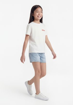 Naeratav tüdruk kõnnib, kandes valget lühikeste varrukatega t-särki punase logoga, heledaid teksapükse ja valgeid tossusid, valgel taustal.