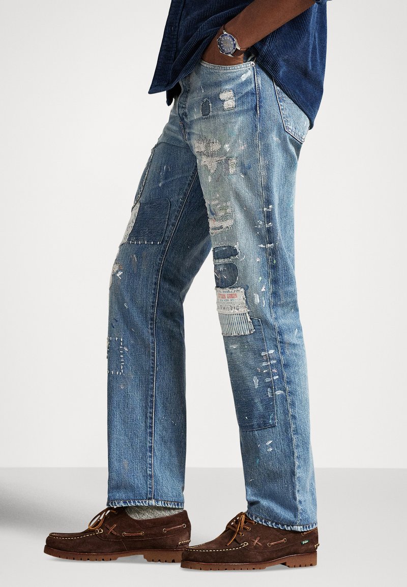 Uomo che indossa jeans blu usurati con toppe e schizzi di vernice, scarpe di camoscio marrone, calzini grigi e una camicia blu con un orologio.