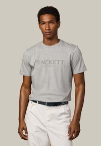 Mand iført en lys grå "Hackett London" T-shirt stoppet ned i hvide bukser med sort bælte foran en ensfarvet beige baggrund.