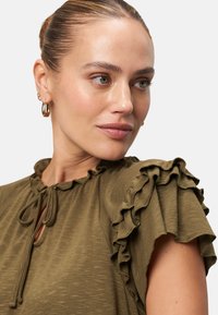Olive green top with ruffled short sleeves, V-neck, and drawstring detail at the neckline. Soft texture with a subtle heathered pattern.