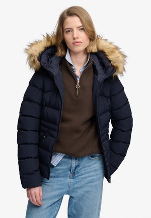 Jeune femme portant une doudoune bleu marine avec capuche en fourrure, un pull marron à fermeture éclair quart, une chemise bleue et un jean bleu clair sur fond blanc.