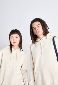 Beiga hoodies med framfickor, dragsnoddar och guld-logotyp. Modellens texturer inkluderar slät bomull och synliga söm detaljer.