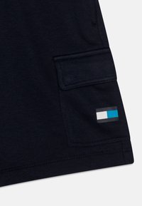 Dunkelblauer Stoff mit einer rechteckigen Tasche, die einen Klappdeckel und ein kleines rechteckiges Logo mit grauen, weißen und blauen Blocks aufweist.