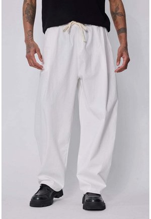 Homme portant un pantalon blanc ample à cordon, des chaussures noires et une chemise noire, avec les bras tatoués le long du corps sur un fond uni.