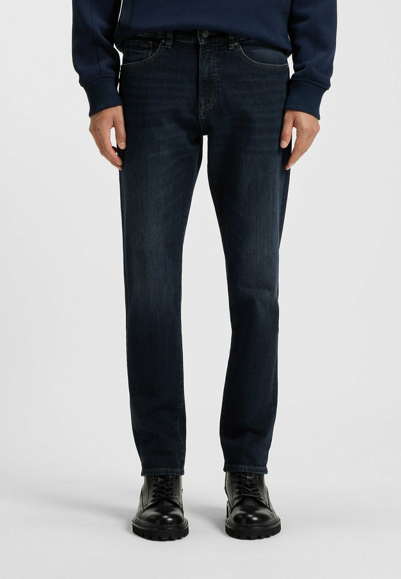 Vaqueros de mezclilla azul oscuro con corte recto, que cuentan con cinco bolsillos y un desgaste sutil. Combinados con botas negras de cordones.