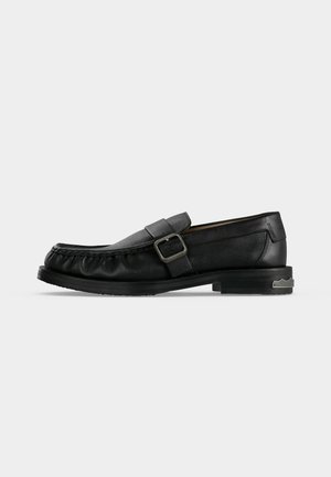 Mocassin en cuir noir avec une bride à boucle, détails cousus autour de la pointe, et un petit talon avec un accent métallique.