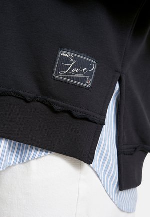 Black sweatshirt hem with "Money or Love?" patch over blue and white striped shirt and white pants.