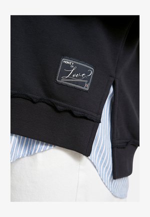 Black sweatshirt hem with "Money or Love?" patch over blue and white striped shirt and white pants.