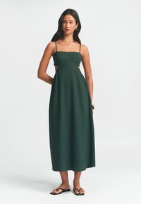 Donkergroene midi-jurk met dunne bandjes, aansluitende corsage, uitsnijdingen aan de zijkanten en een vloeiende rok. Gemaakt van een gestructureerde stof. Eenvoudig ontwerp.