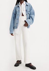 Giacca in denim azzurro chiaro con vestibilità comoda, camicia bianca con bottoni, pantaloni bianchi a gamba larga e mocassini neri, con una finitura texture.