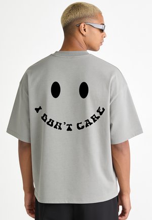 Personne aux cheveux blonds courts portant des lunettes de soleil noires et un t-shirt gris avec un visage souriant noir et le texte "I DON'T CARE" dans le dos.