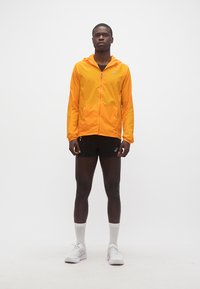 Άνδρας μοντέλο όρθιος, φοράει φωτεινό πορτοκαλί μπουφάν με κουκούλα, μαύρο αθλητικό σόρτς, λευκές κάλτσες μέχρι τον αστράγαλο και λευκά παπούτσια για τρέξιμο.