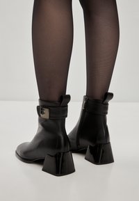Botine negre din piele cu tobă, cu toc pătrat, închidere cu fermoar și curea cu accent metalic. Designul este elegant și modern.