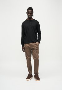 Chemise henley noire à manches longues, pantalon beige et baskets marron. Tissu lisse, couleurs unies, coupe décontractée et boutons sur la chemise.