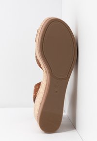 Brązowa sandał z teksturowaną, warstwową podeszwą, ozdobiony złotymi ćwiekami, z widoczną marką "GUESS" i rozmiarem "37" na spodzie.