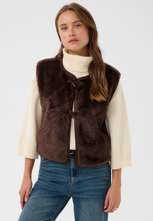 Femme portant une veste en peluche marron foncé par-dessus un pull à col roulé blanc et un jean bleu, debout devant un fond clair uni.