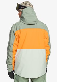 Chaqueta resistente al agua en paneles verdes, naranjas y blancos. Cuenta con capucha y puños elásticos, diseñada para actividades en climas fríos.