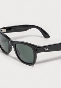Sorte plastiks solbriller med grønne tonede linser, rektangulær form og et synligt kamera på siden. Ray-Ban logo på stængerne.
