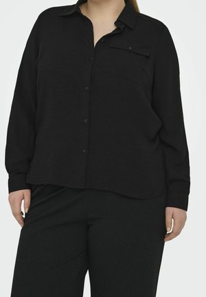 Kvinde iført en løs sort skjorte med knapper og lange ærmer samt matchende sorte bukser mod en ensfarvet baggrund.