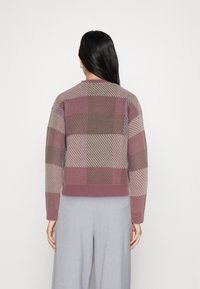 Pull en maille avec un motif à carreaux dans des nuances de rose, marron et gris. Ourlet et poignets côtelés. Le modèle porte un pantalon gris.