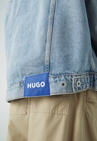 Giacca di jeans blu chiaro con un'etichetta blu a contrasto con la scritta "HUGO". Bottoni a scatto visibili e un primo piano della texture del tessuto.