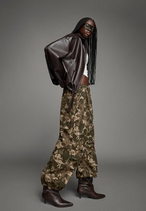 Braune Leder-Oversized-Jacke, weißes Crop-Top, grüne Camouflage-baggy-Hosen und braune Stiefeletten mit Absatz. Zubehör umfasst große grüne Sonnenbrillen.