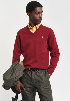 Mann trägt einen roten V-Ausschnitt-Pullover über einem gelb gestreiften Hemd, graue Hose, hält eine graue Jacke und eine schwarze Tasche, schaut nach rechts.