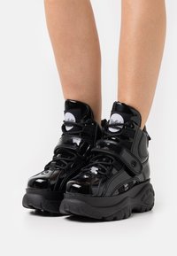 Black, glossy high-top sneakers with a strap, thick rubber sole, and textured accents. Features logo on tongue and black laces.