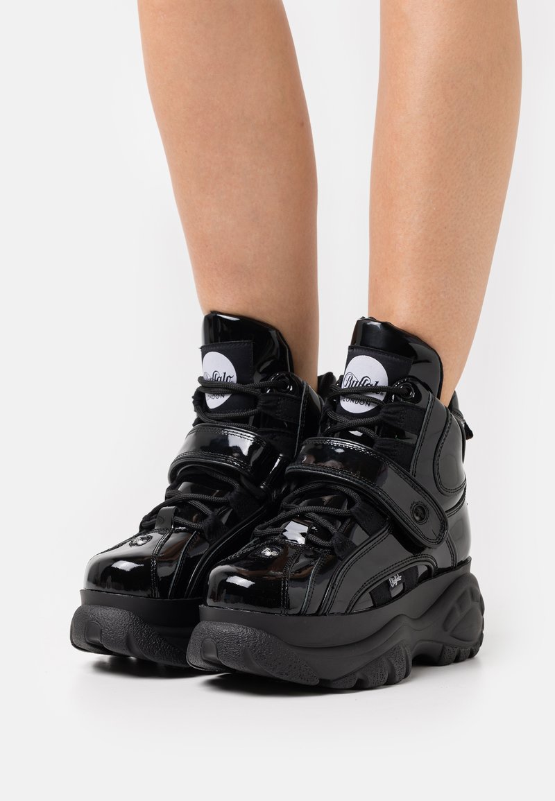Black, glossy high-top sneakers with a strap, thick rubber sole, and textured accents. Features logo on tongue and black laces.