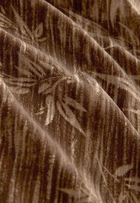 Strukturierter Stoff in einem satten Braunton, mit einem subtilen Blumenmuster und einer weichen, samtigen Optik. Falten bilden dynamische Bögen.