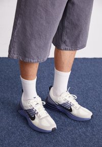 Vita och marinblå atletiska sneakers med mesh-överdel, utformade med en svart swoosh, lila mellansula och vita snören. Bärs med vita ankelsockor.