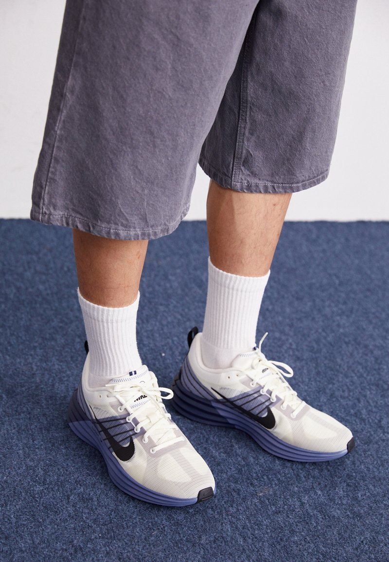 Vita och marinblå atletiska sneakers med mesh-överdel, utformade med en svart swoosh, lila mellansula och vita snören. Bärs med vita ankelsockor.