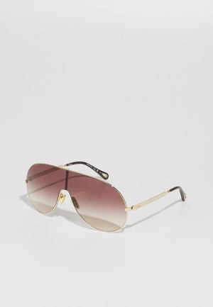 Gafas de sol aviador doradas con lentes marrones degradados, montura de metal fina y acentos negros en las patillas, con un logo en un lado.
